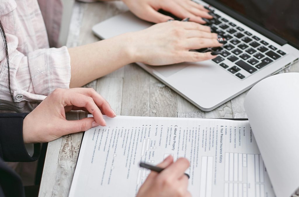 Close-up of two professionals reviewing a detailed document, with one pointing at the text while the other types on a laptop, highlighting collaborative document analysis and preparation.