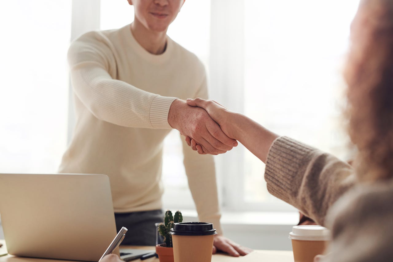 Two professionals shaking hands over a desk in a bright office setting, symbolizing a successful business agreement or partnership.